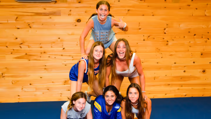 Six smiling girls forming a human pyramid on a blue mat against a wooden wall background.