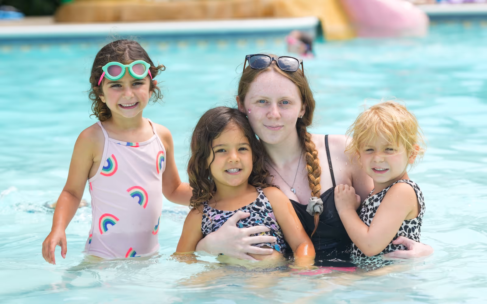 Young woman with braided hair and sunglasses on her head in a pool with three smiling children wearing swimsuits.