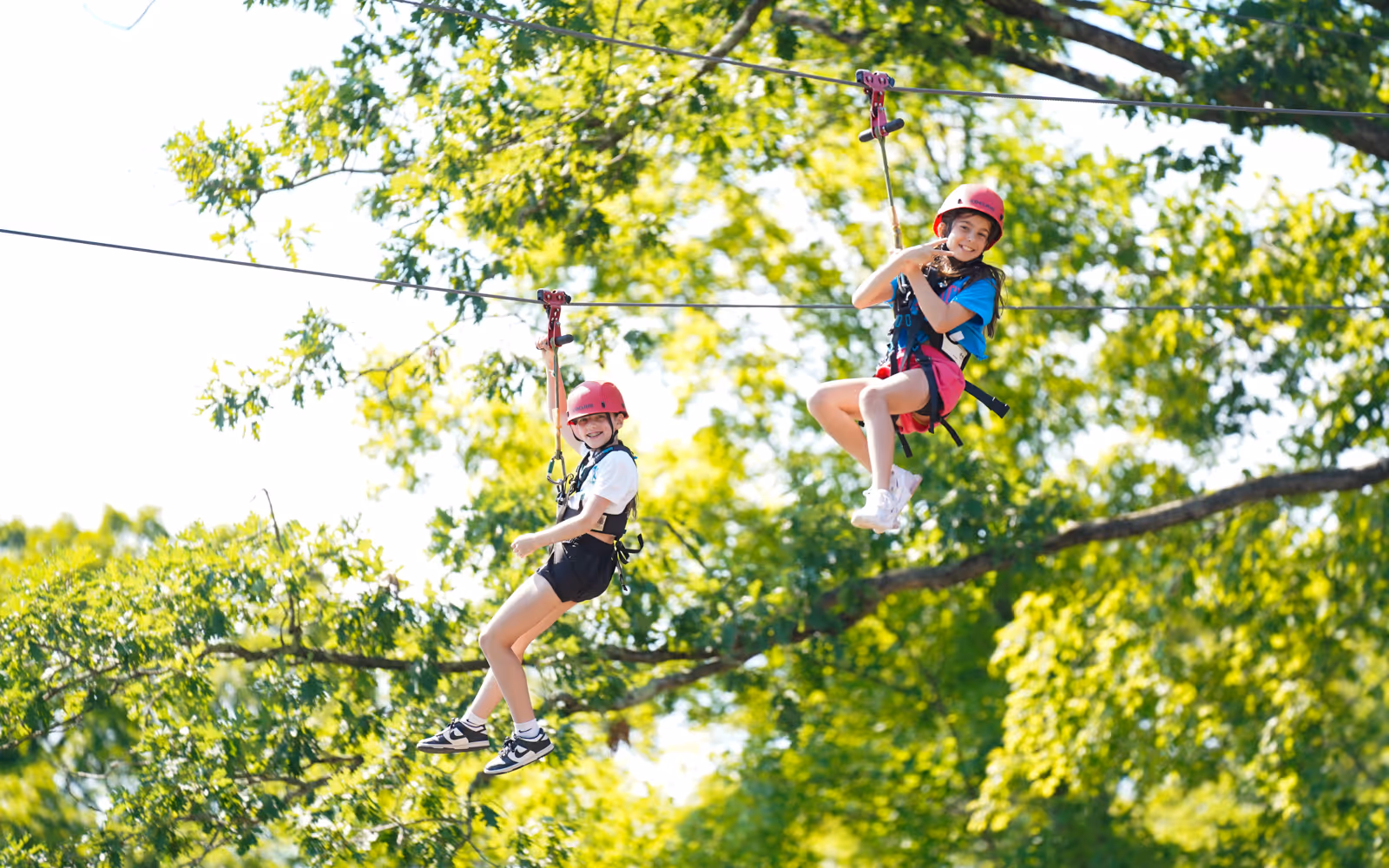 Two girls wearing helmets and harnesses smiling while riding a zipline through green trees on a sunny day.
