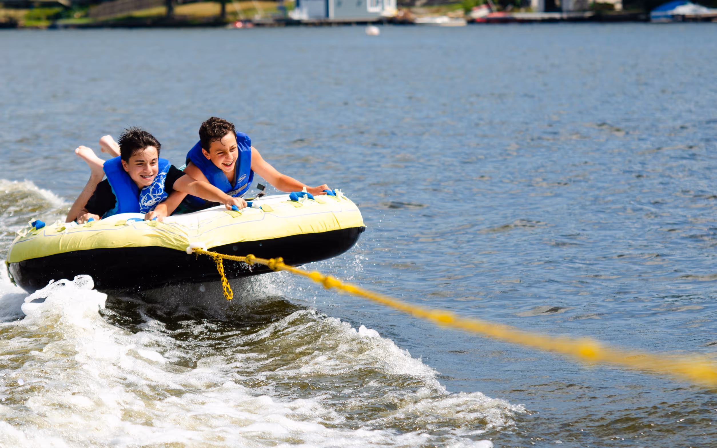 Two boys wearing blue life jackets smiling and holding onto a yellow inflatable tube being pulled across water.