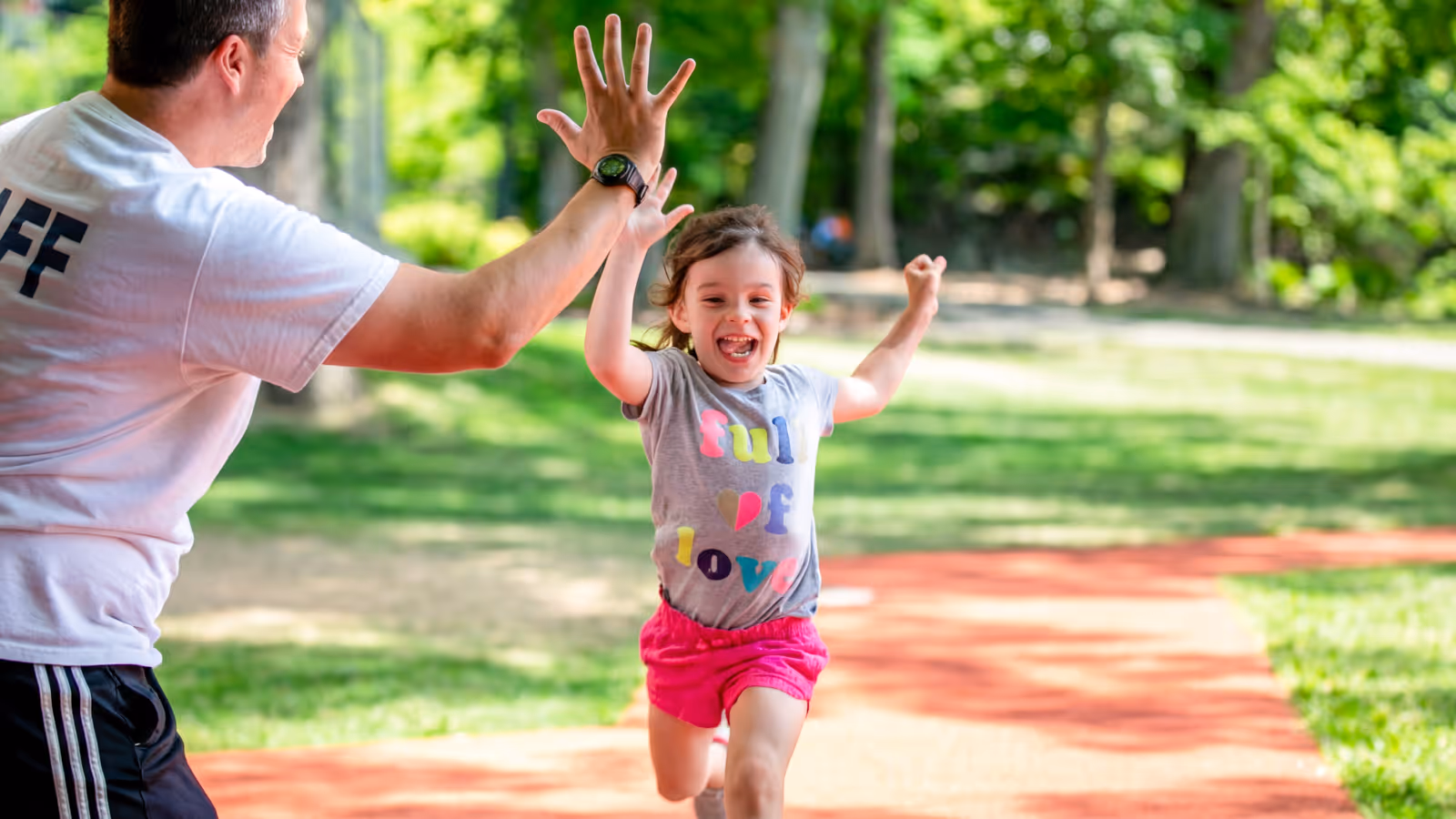Young girl happily running outdoors and giving a high five to a man wearing a white shirt labeled 'STAFF'.