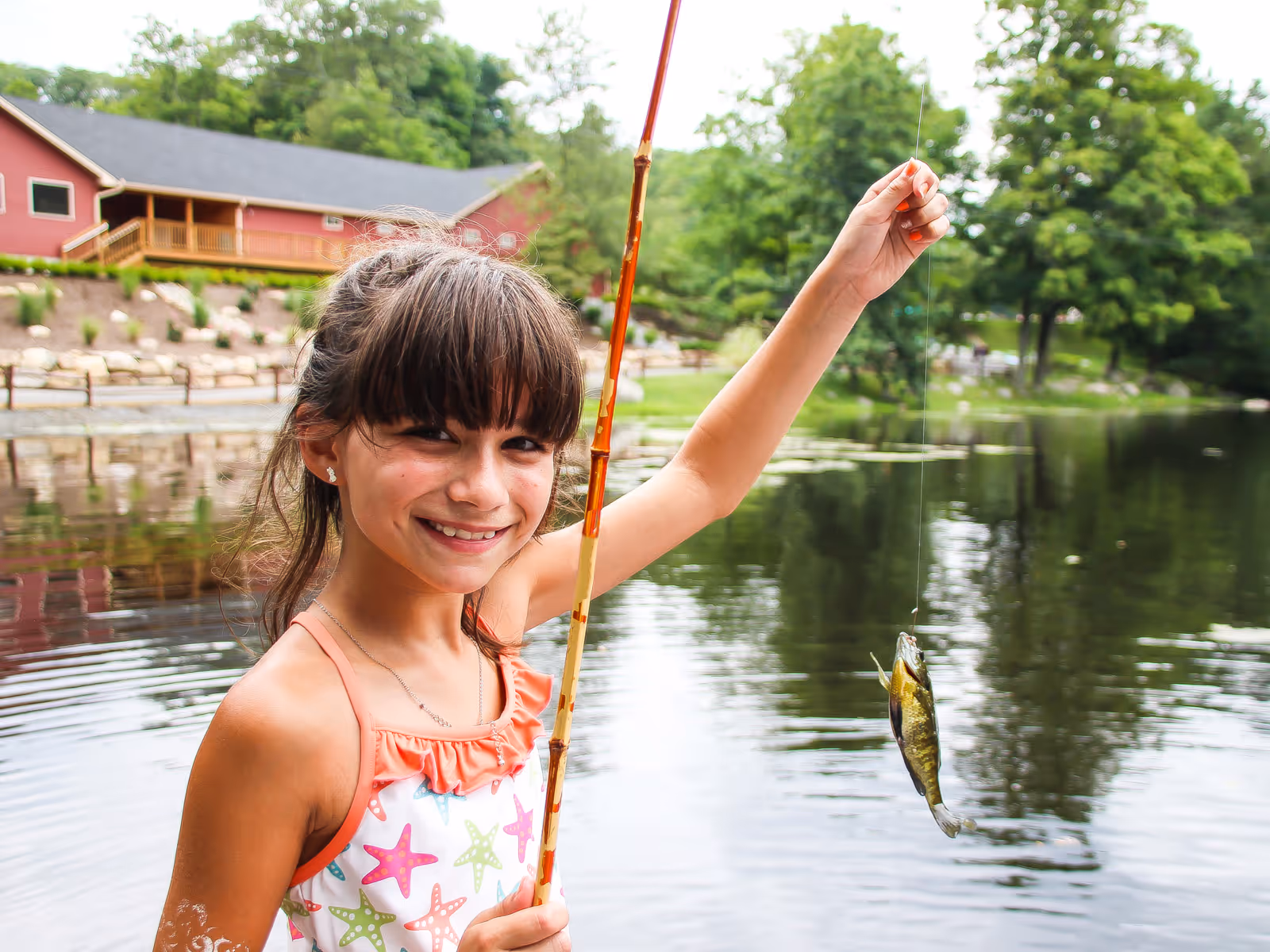 Smiling girl in a starfish-patterned swimsuit holding a fishing rod with a small fish caught on the line by a lake.