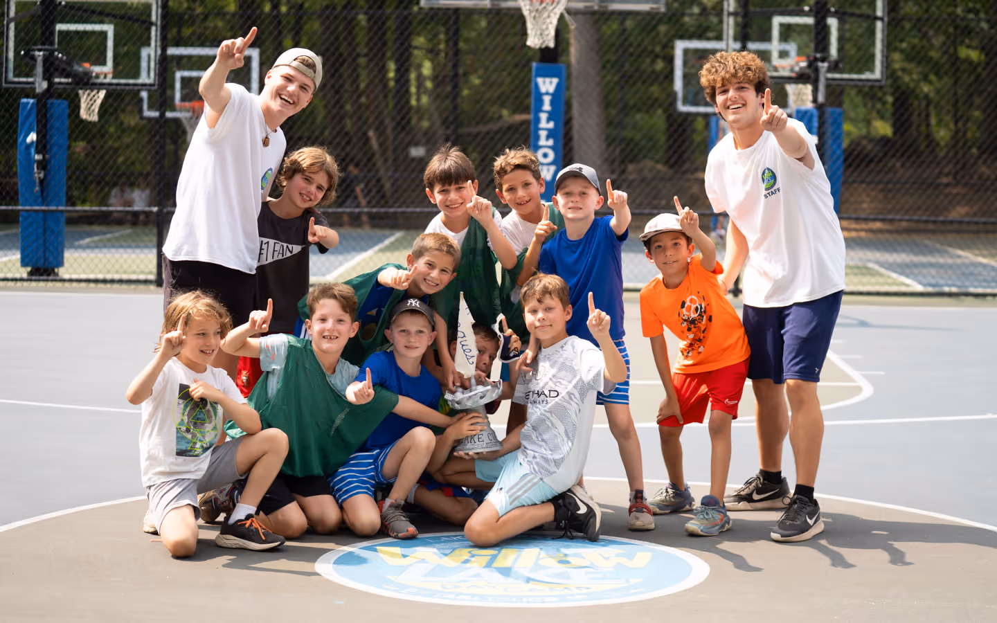 Group of boys and two camp counselors on a basketball court, smiling and holding a trophy while showing number one with their fingers.