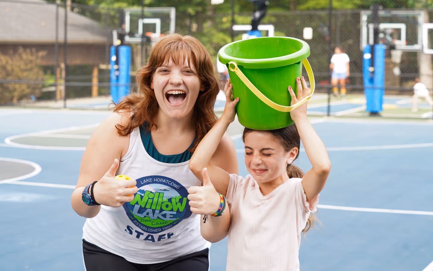 Smiling camp counselor giving thumbs up beside a girl holding a green bucket on her head at an outdoor basketball court.