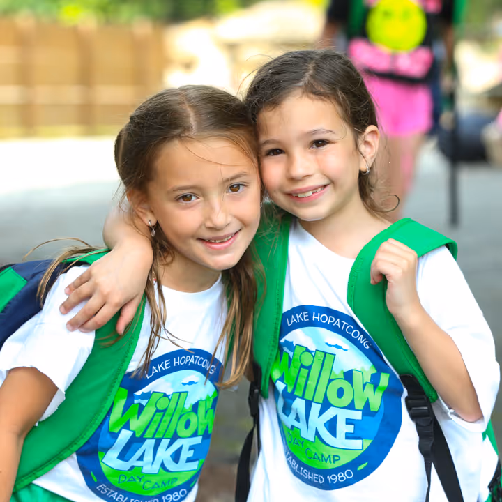 Two smiling girls wearing white Willow Lake Day Camp shirts and green backpacks with arms around each other.