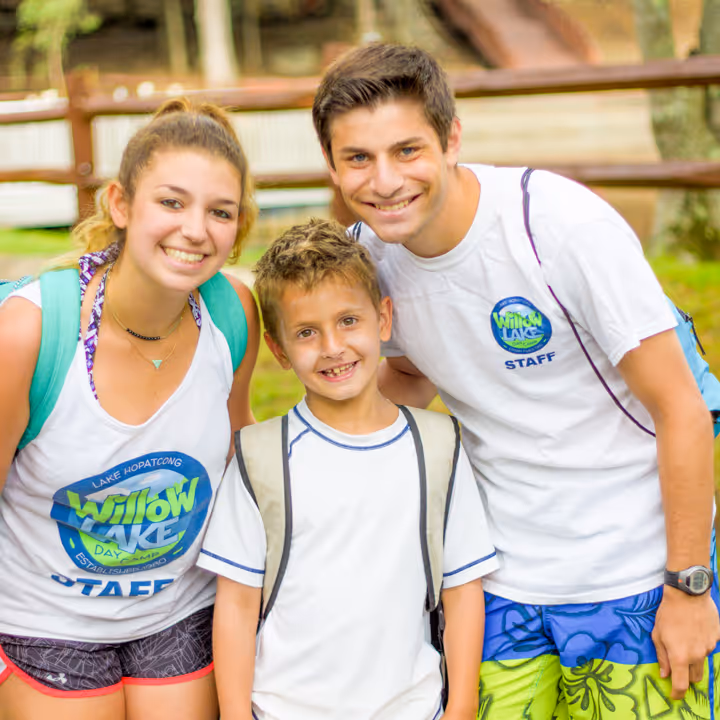 Two camp staff members smiling with a young camper wearing backpacks at an outdoor camp setting.