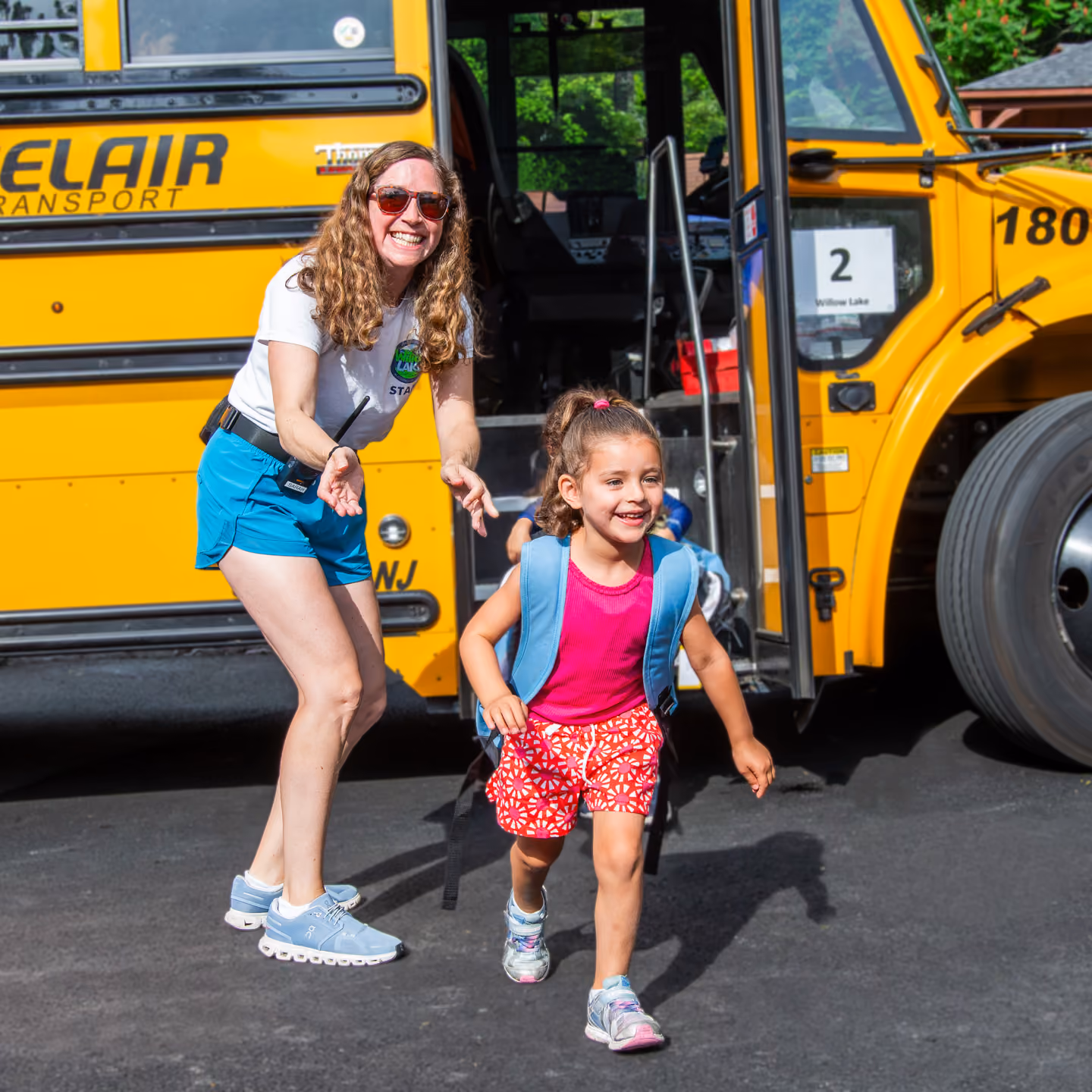 Smiling female counselor with sunglasses gestures as young girl with backpack runs from a yellow school bus.