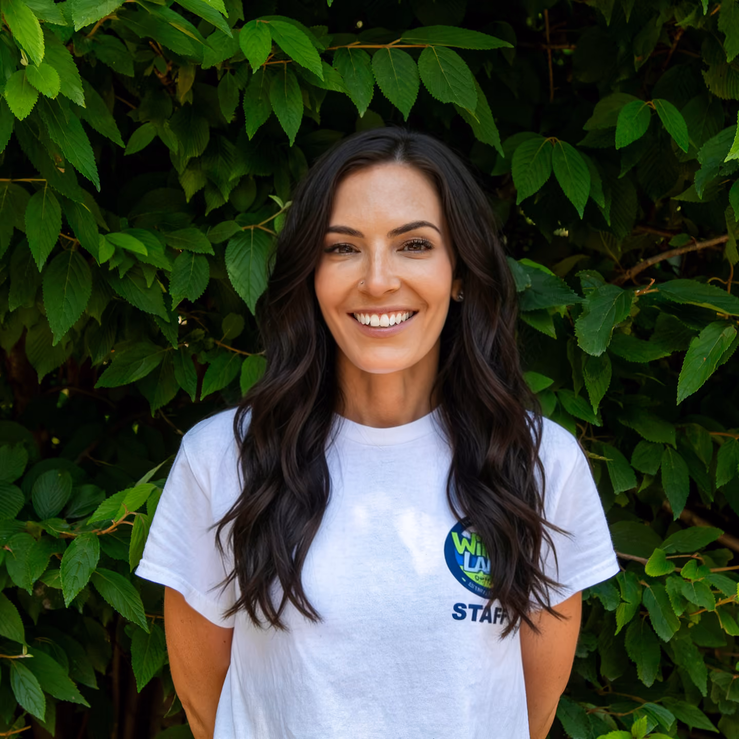 Alena Hardison wearing a white staff shirt standing in front of green leaves.