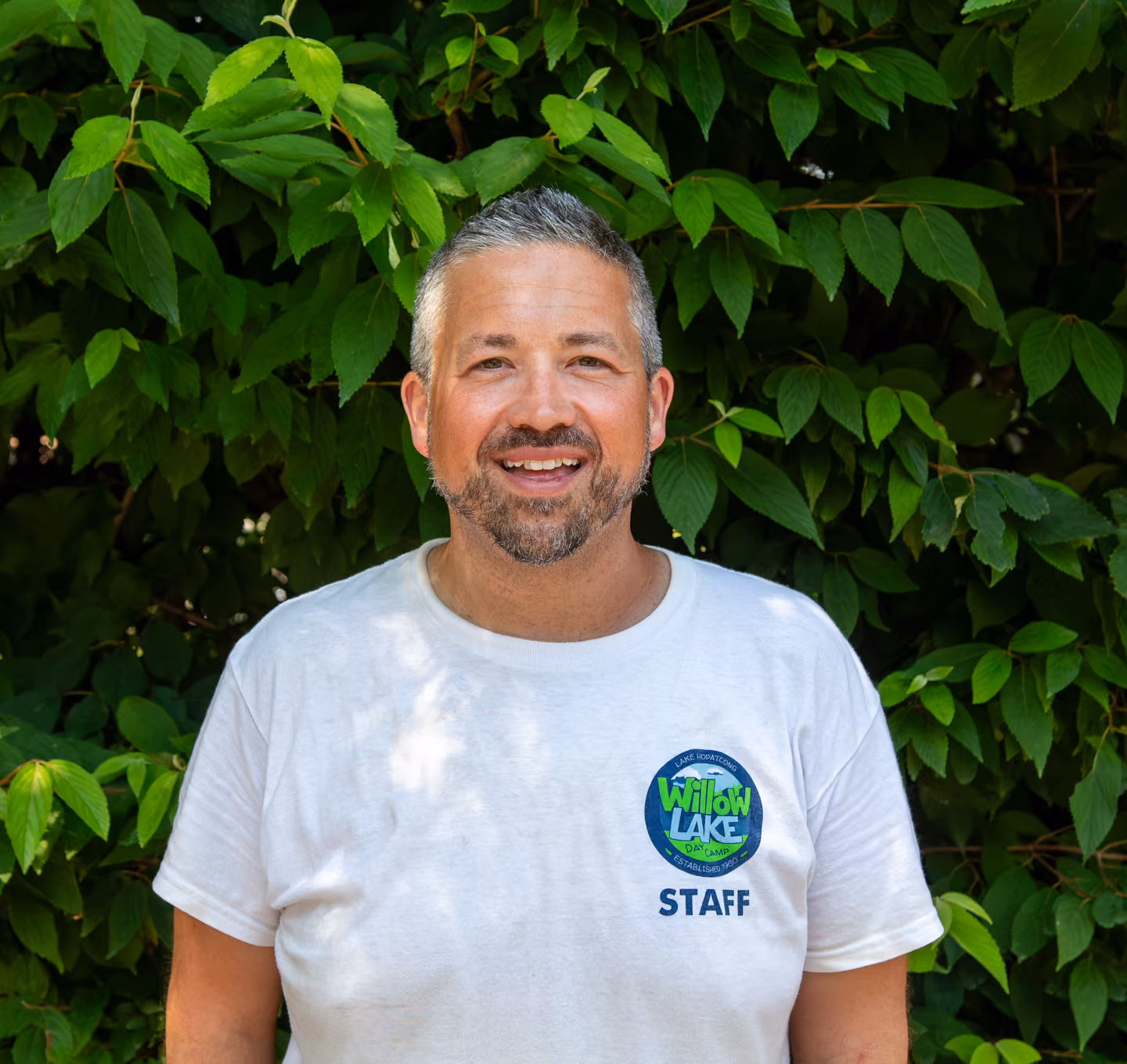 Zach Handler wearing a white Willow Lake Day Camp staff shirt standing in front of green leafy bushes.