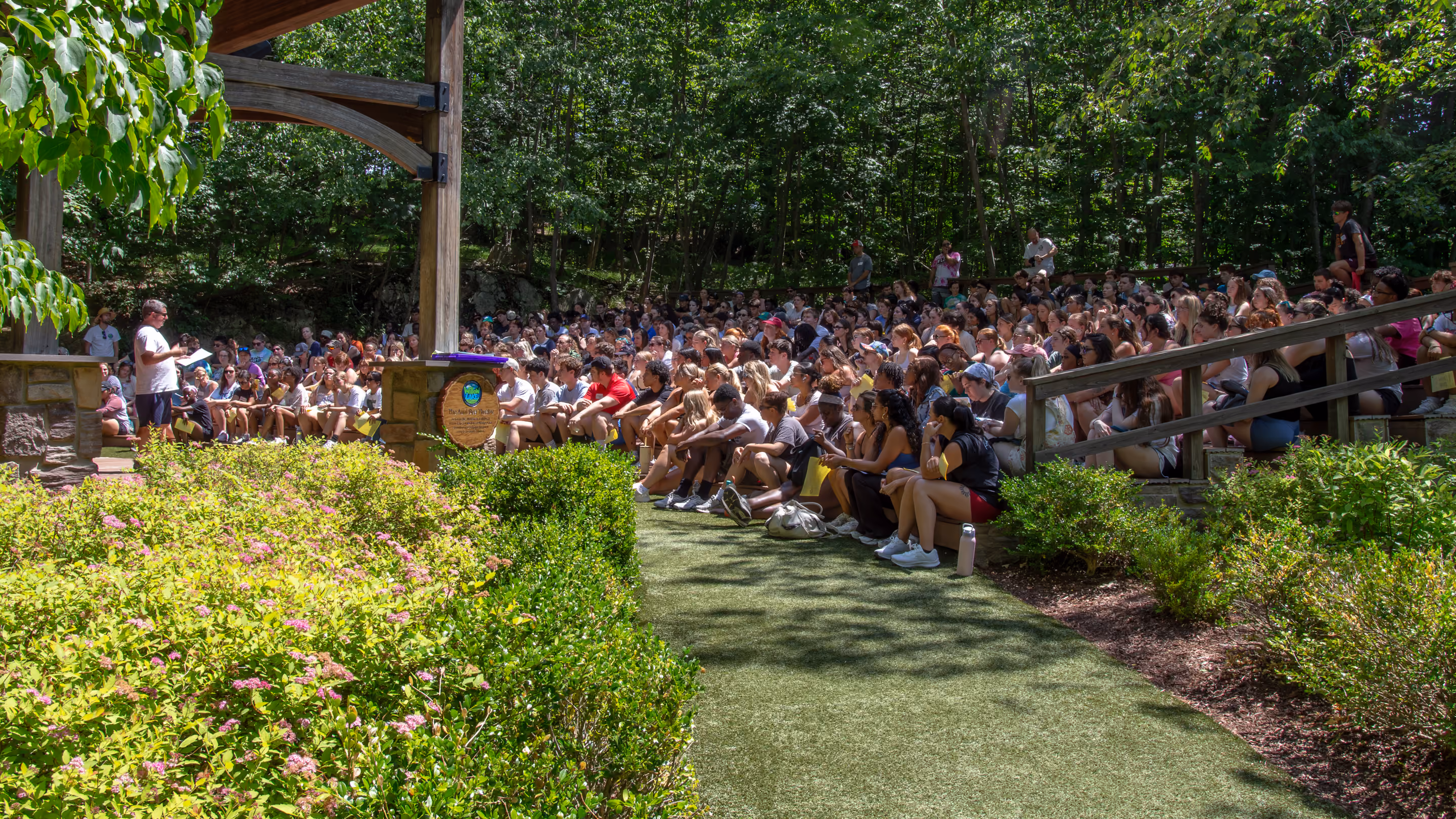 Man speaking with a microphone to a large outdoor audience of children seated on wooden bleachers surrounded by trees.