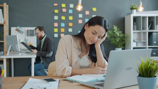 Tired young woman is sleeping in office then waking up and working with laptop in office, colleague is busy in background. People and lifestyle concept.