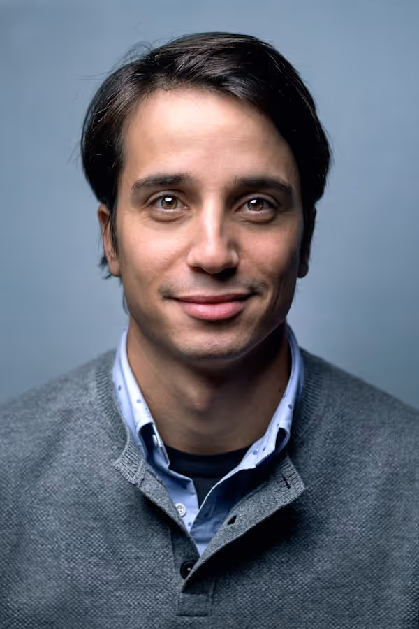 Portrait of a smiling man with dark hair wearing a grey sweater over a collared shirt against a plain blue background.