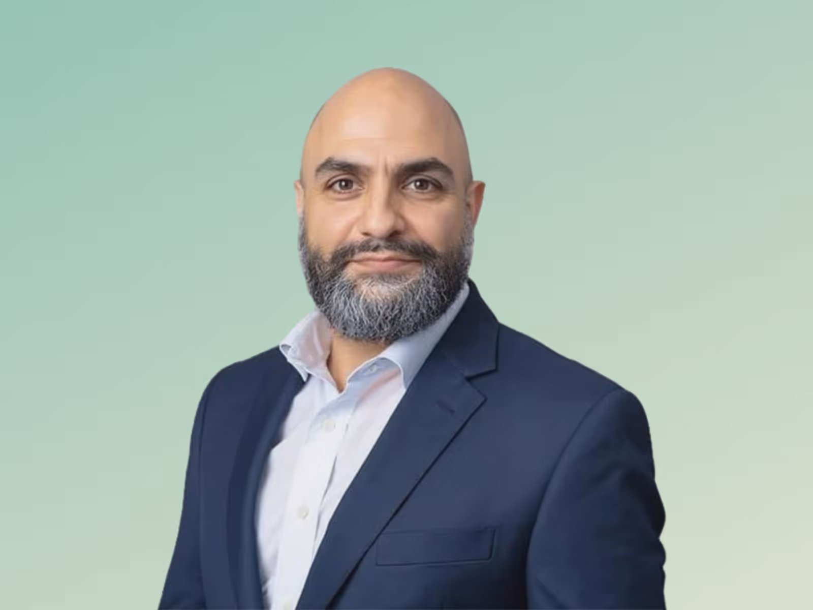 Portrait of a bald man with a graying beard wearing a navy suit jacket and white shirt against a green gradient background.