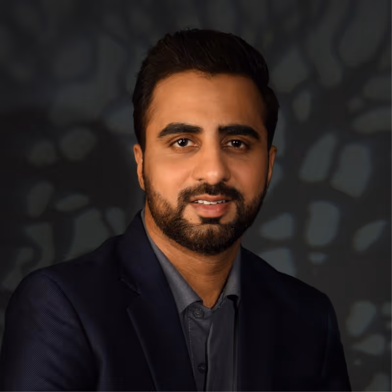 Portrait of a man with a beard wearing a dark blazer and shirt against a softly patterned dark background.