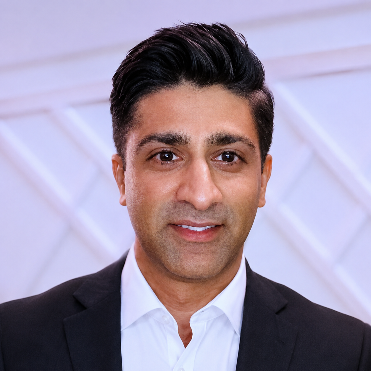 Portrait of a man with dark hair wearing a black suit and white shirt at an indoor event.