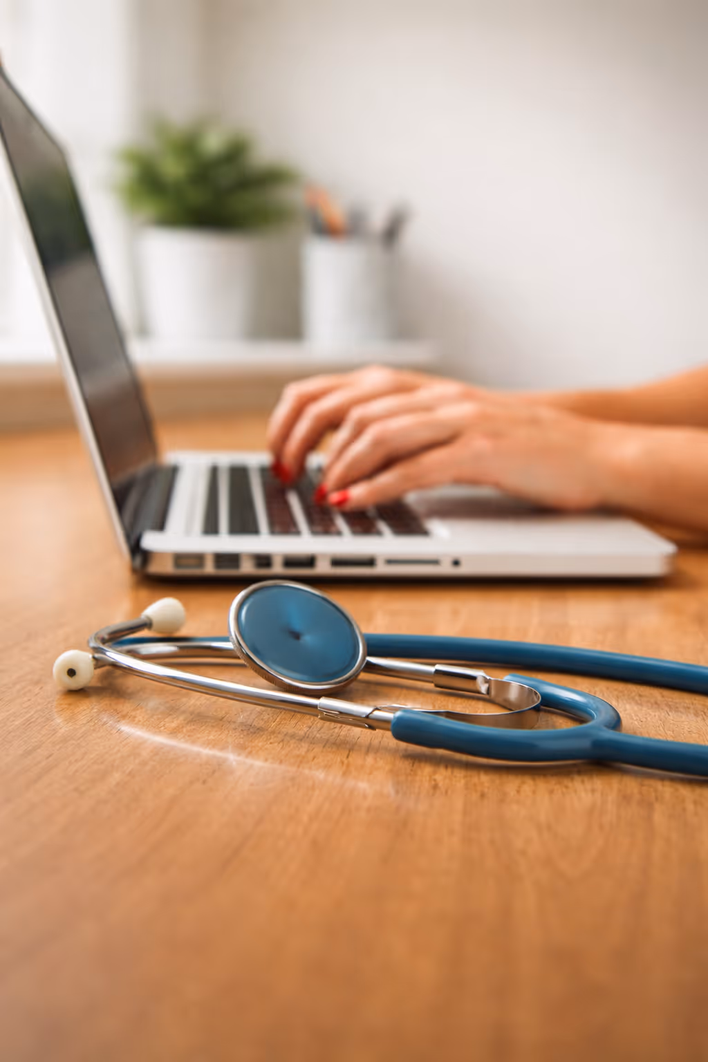 Blue stethoscope on a wooden desk with hands typing on a laptop keyboard in the background.