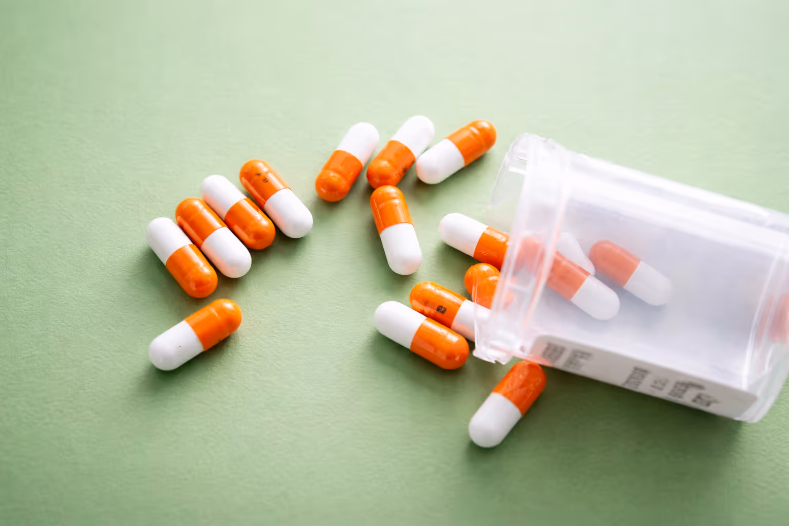 Orange and white capsules spilling out of a clear prescription pill bottle on a green surface.