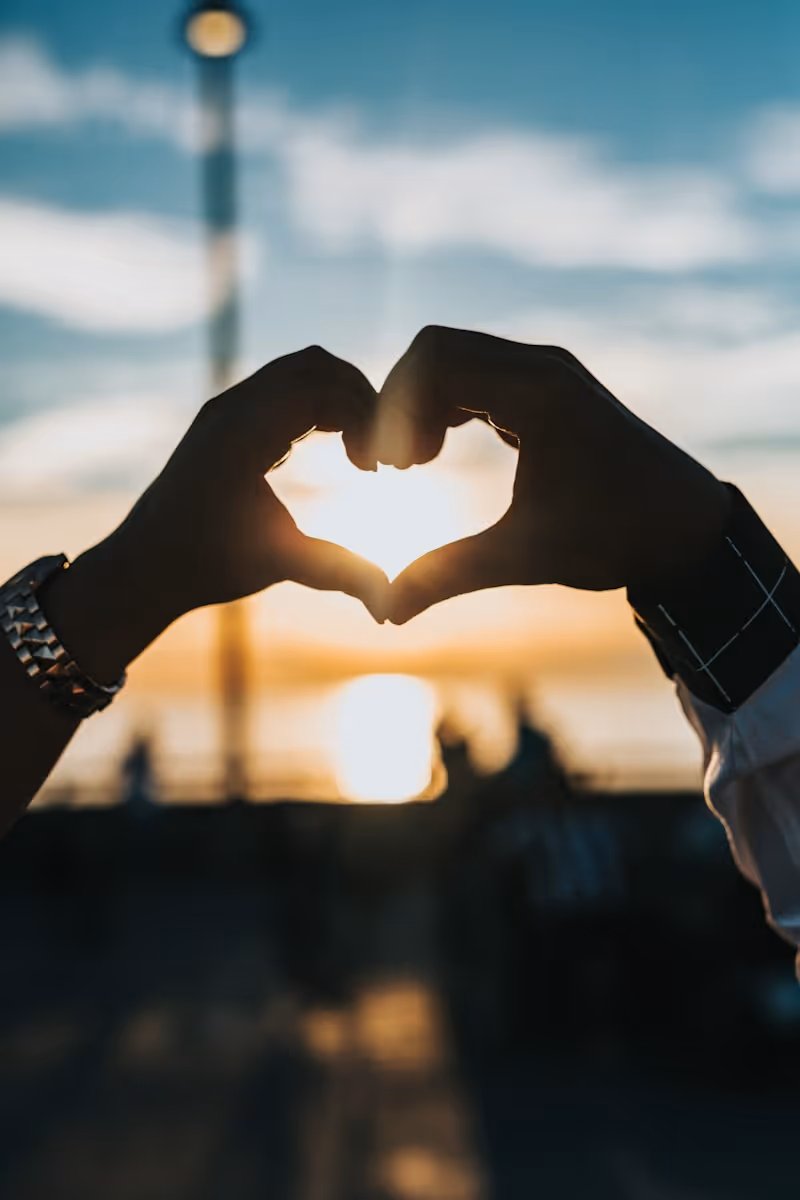 Two hands forming a heart shape with the setting sun visible through the heart.