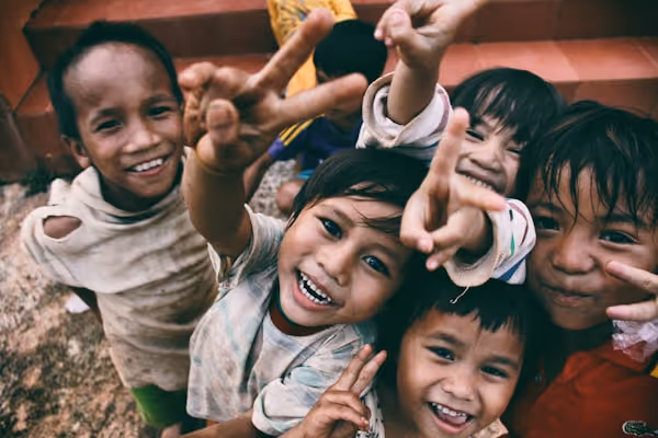 A group of joyful children smiling and making peace signs with their hands while looking up at the camera.
