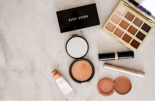 Collection of makeup products including a Bobbi Brown eyeshadow palette, compact powder, lipstick, mascara, blush, and a small tube on a marble surface.