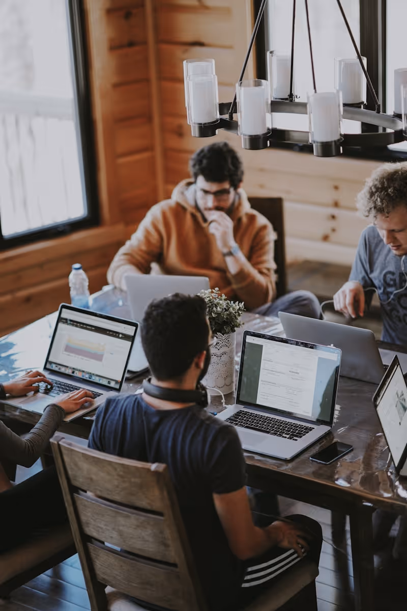 Four people working on laptops around a wooden table in a cozy room with wooden walls and a chandelier.