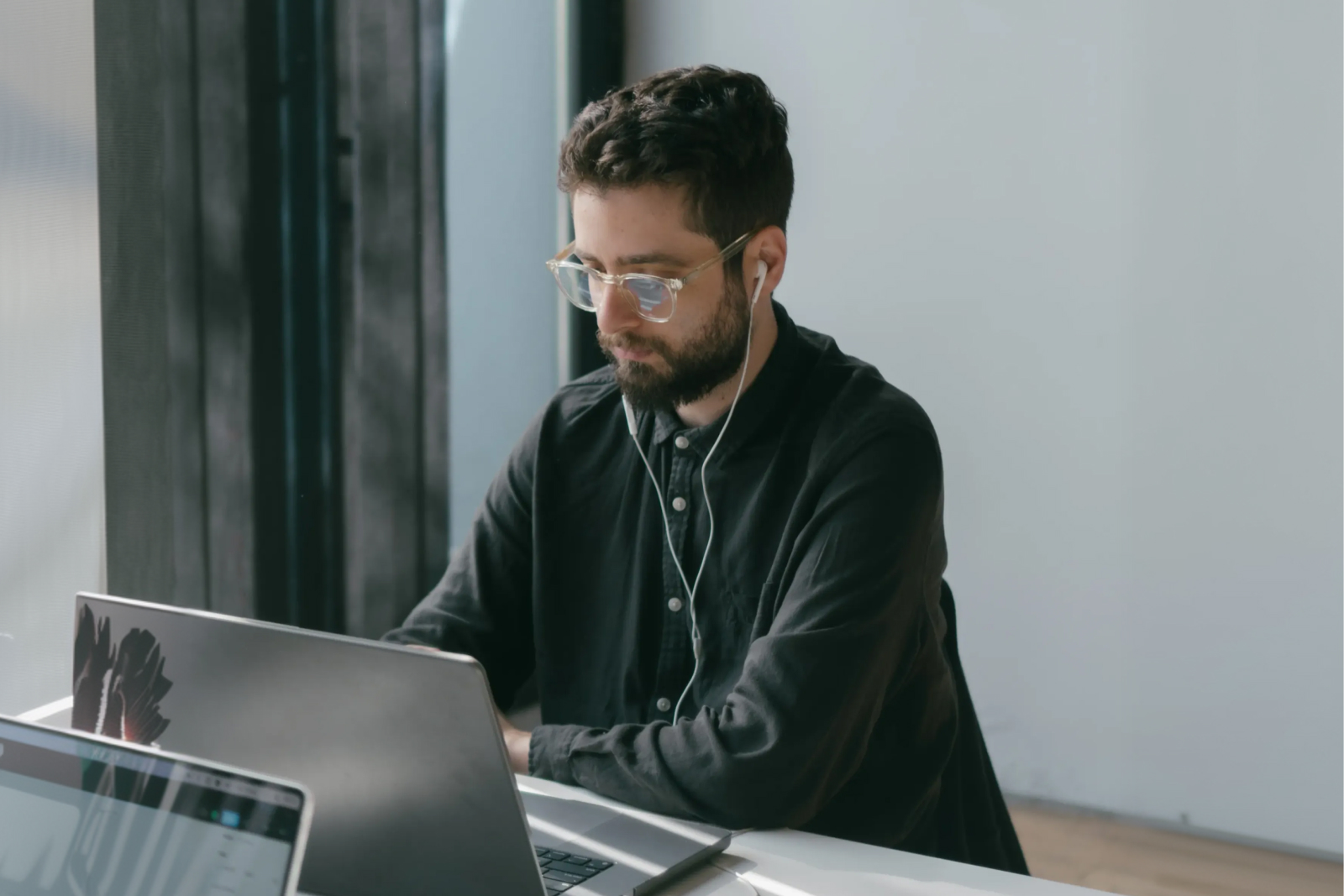 Man wearing clear glasses and earphones working on a laptop at a desk near a window.