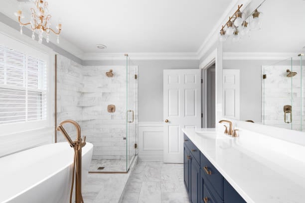 Bright modern bathroom with a white freestanding tub, glass shower, blue cabinetry, and gold fixtures.
