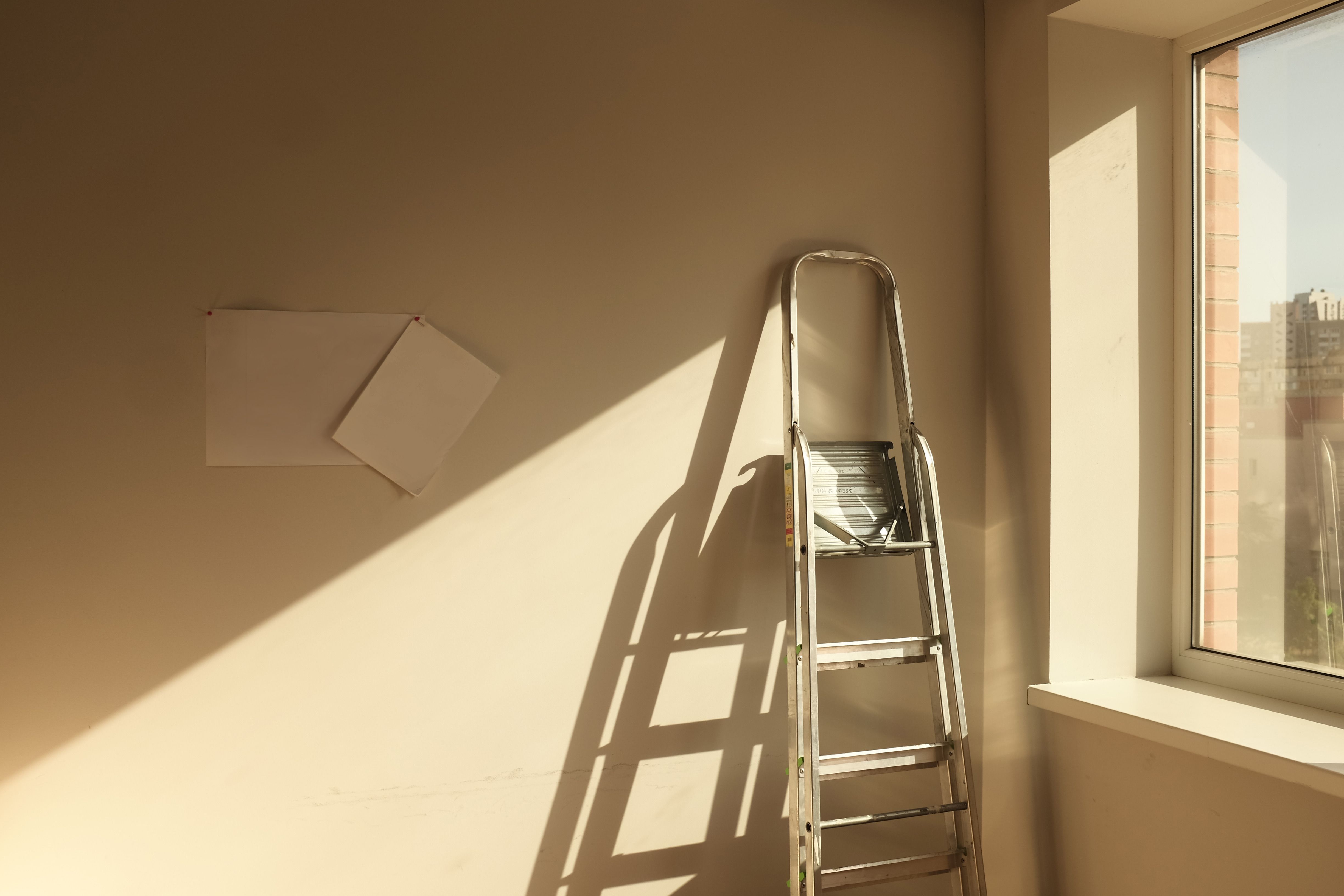 Metal ladder leaning against wall near window with sunlight casting shadows inside a room.