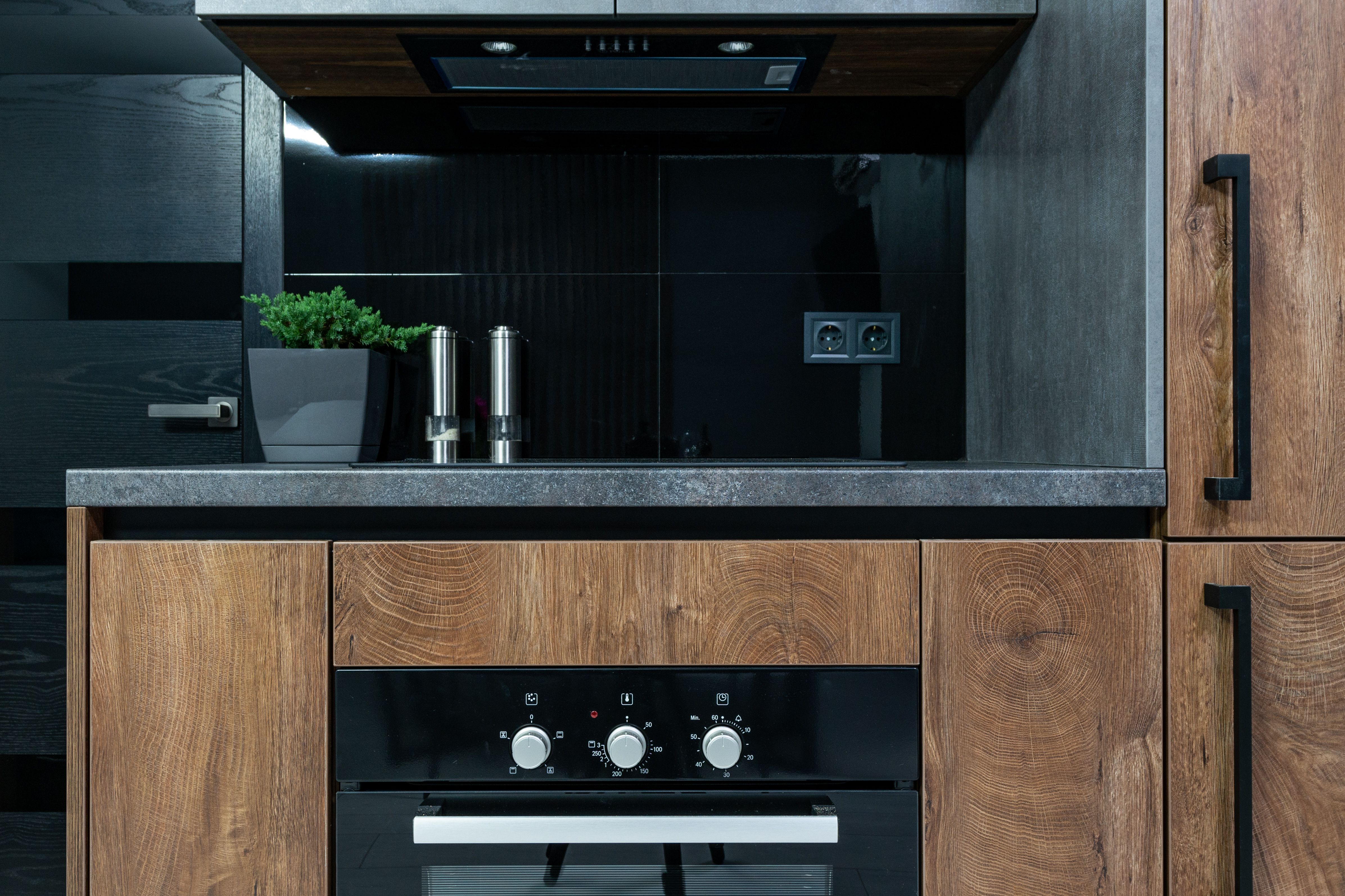 Modern kitchen oven with black panel and wooden cabinets under a gray countertop with pepper mills and green plant.