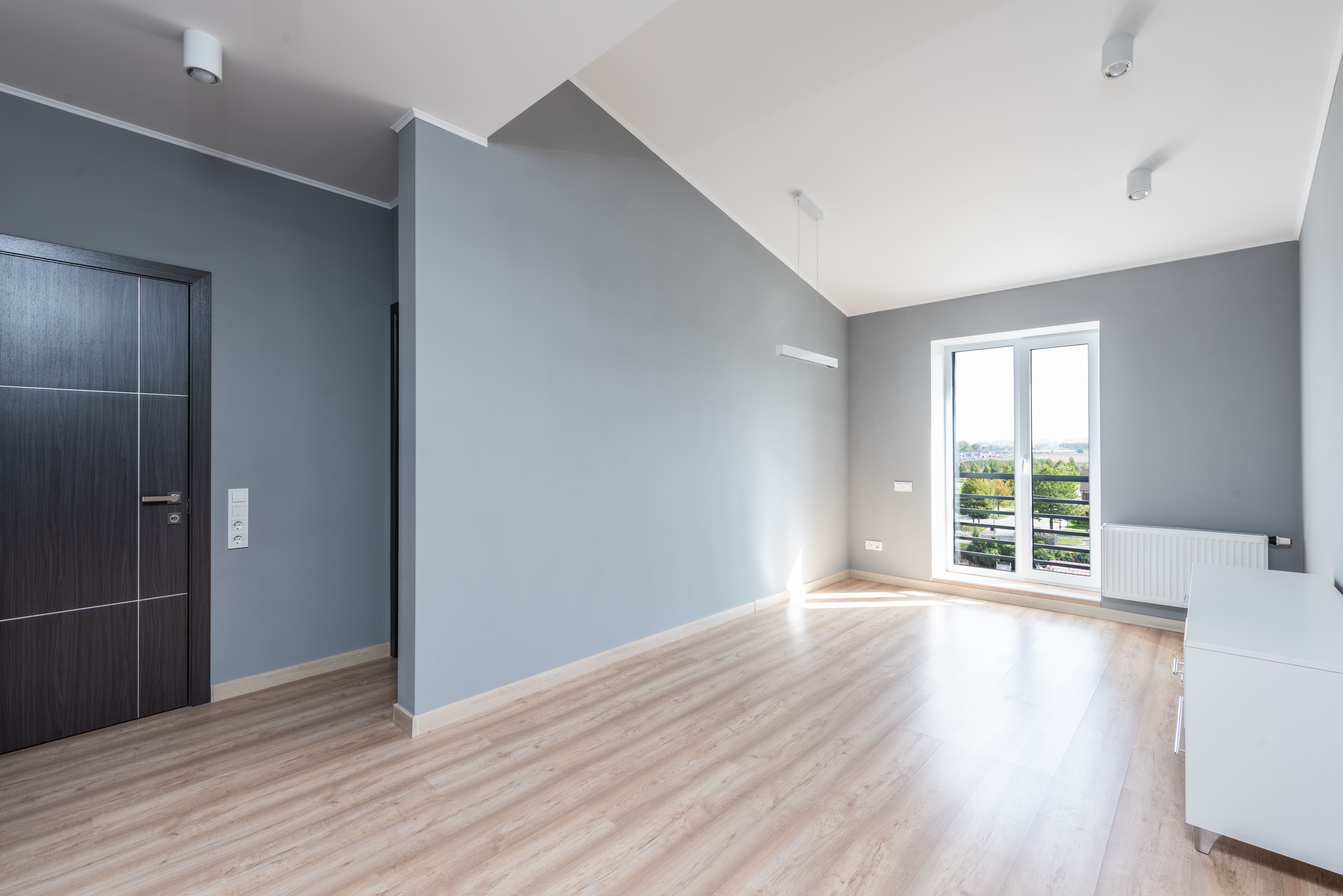 Empty modern room with light wood floor, gray walls, dark door, and large window with balcony.