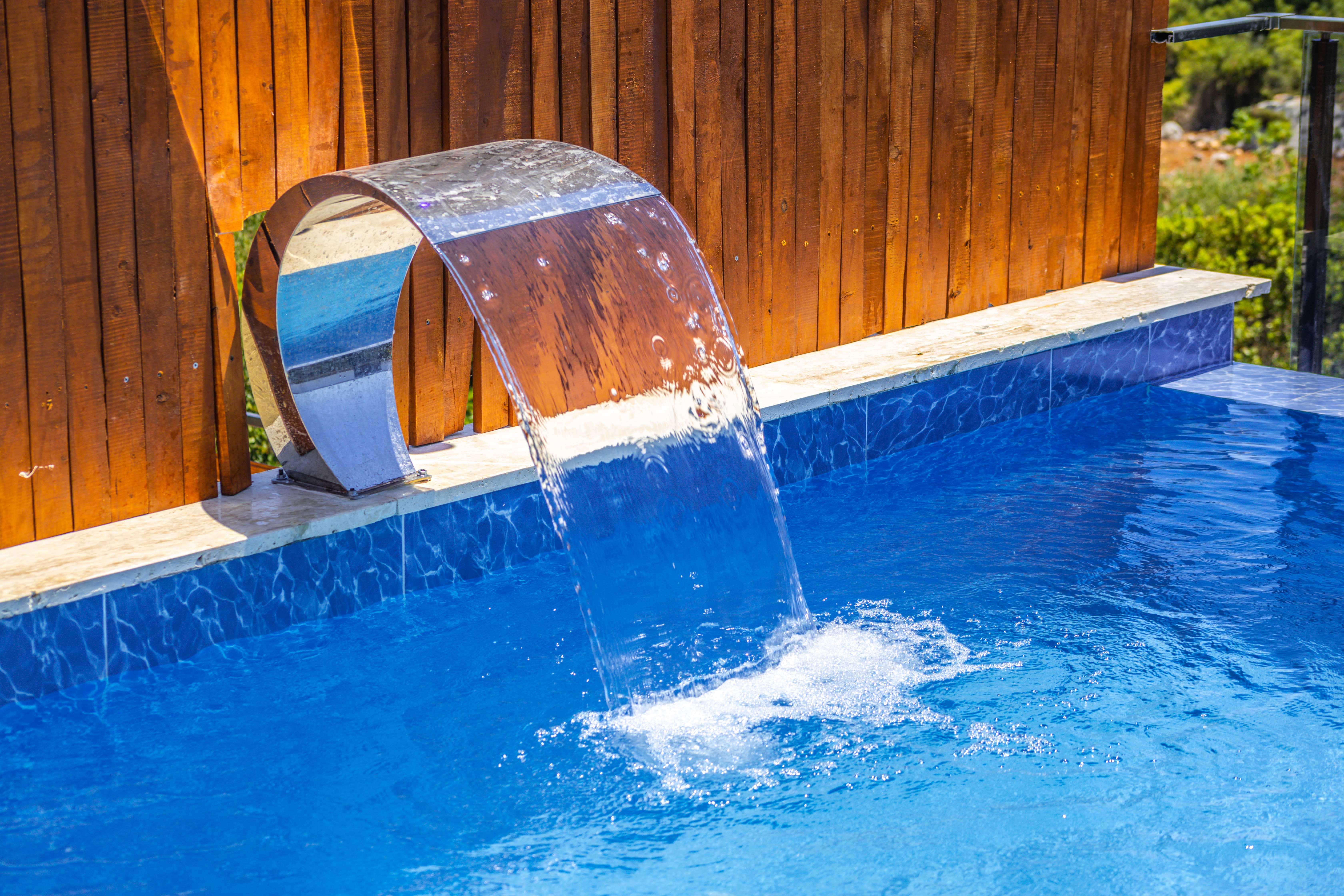 Curved metal water spout flowing water into a blue tiled outdoor swimming pool.