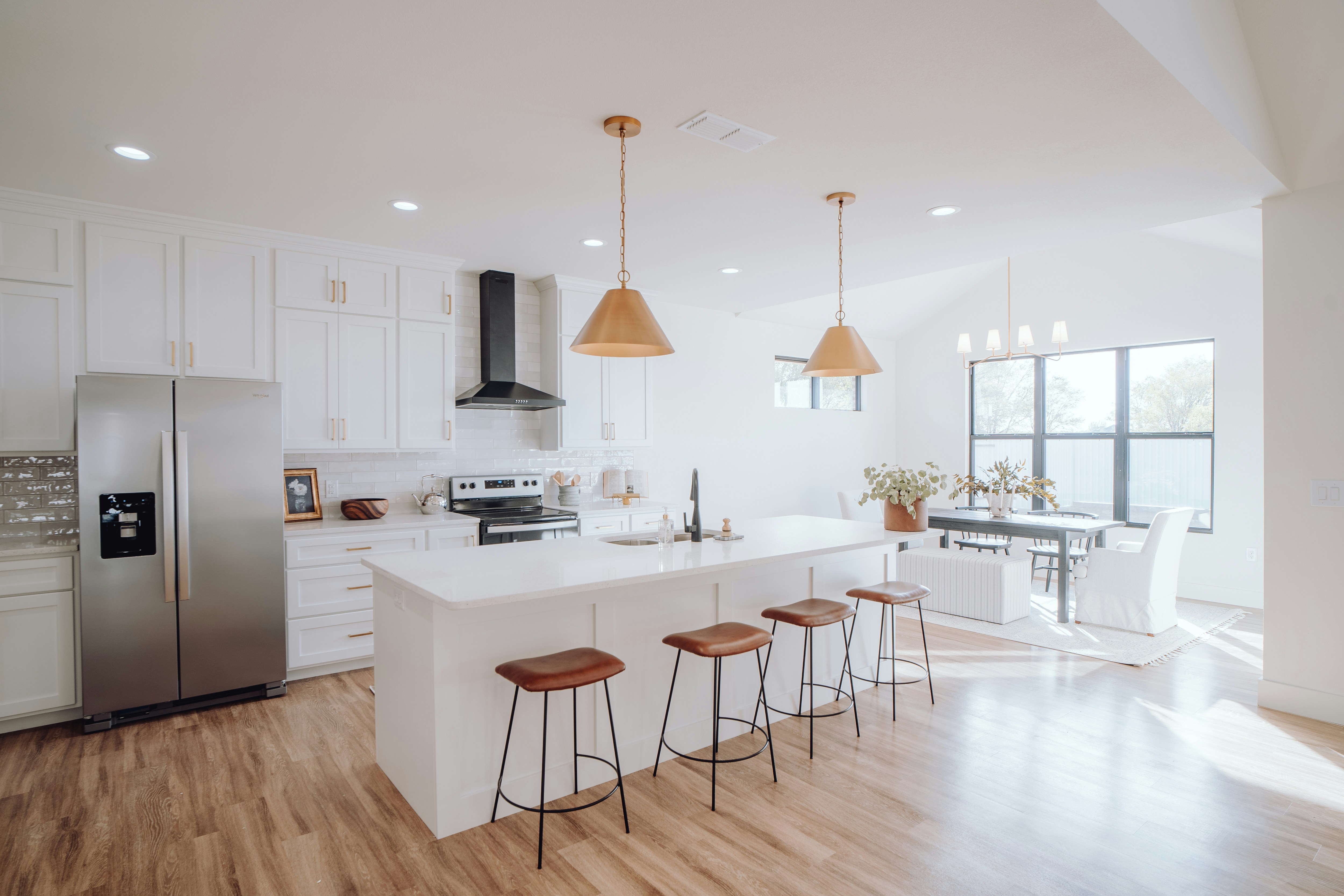Bright kitchen with white island, four stools, stainless fridge, and dining table by large windows.