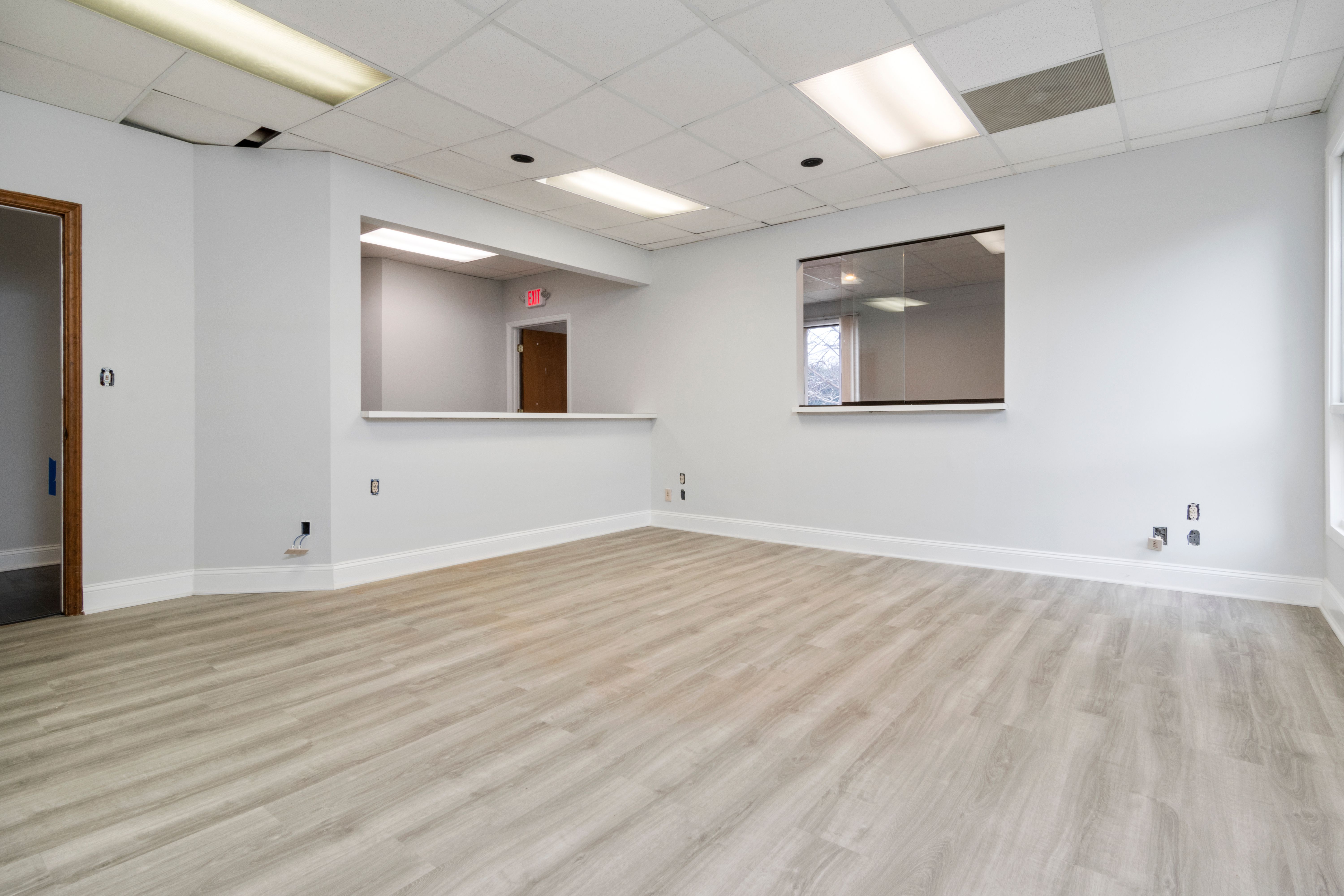 Empty room with light gray walls, large window opening, wood trim door, and light wood flooring.