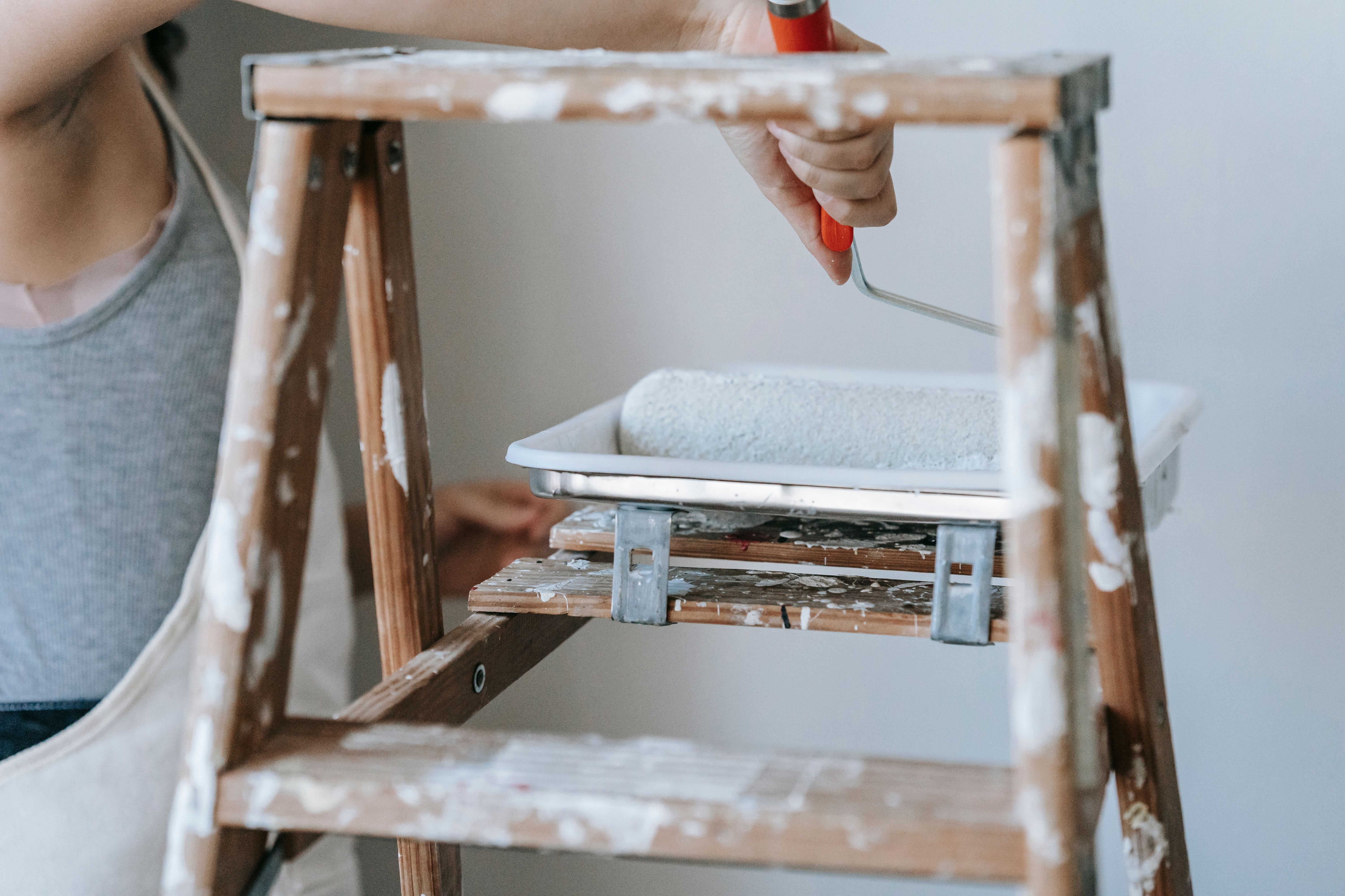 Hand dipping paint roller with red handle into paint tray on a paint-splattered wooden ladder.