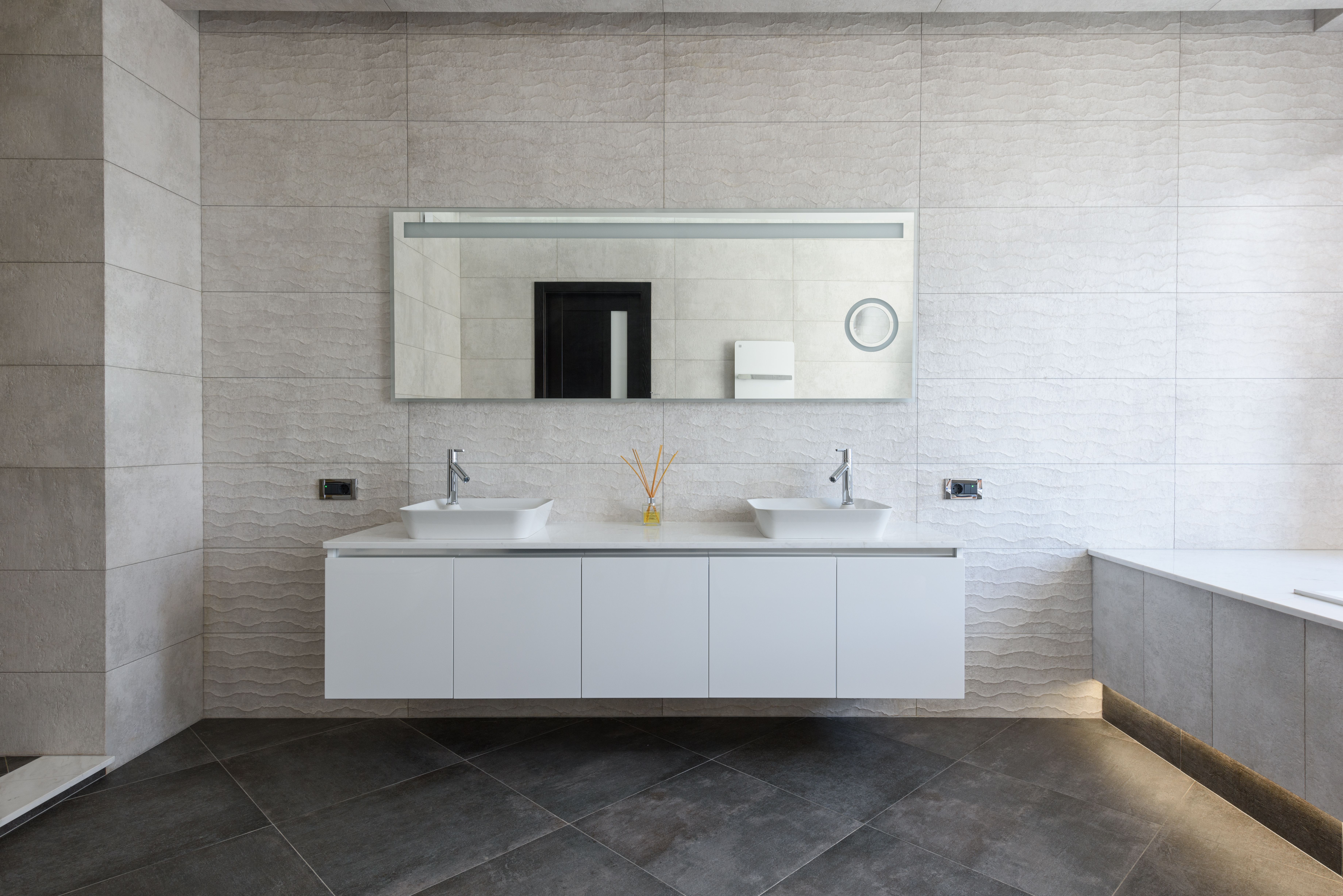 Modern bathroom with two white vessel sinks, large mirror, textured walls, and dark floor tiles.