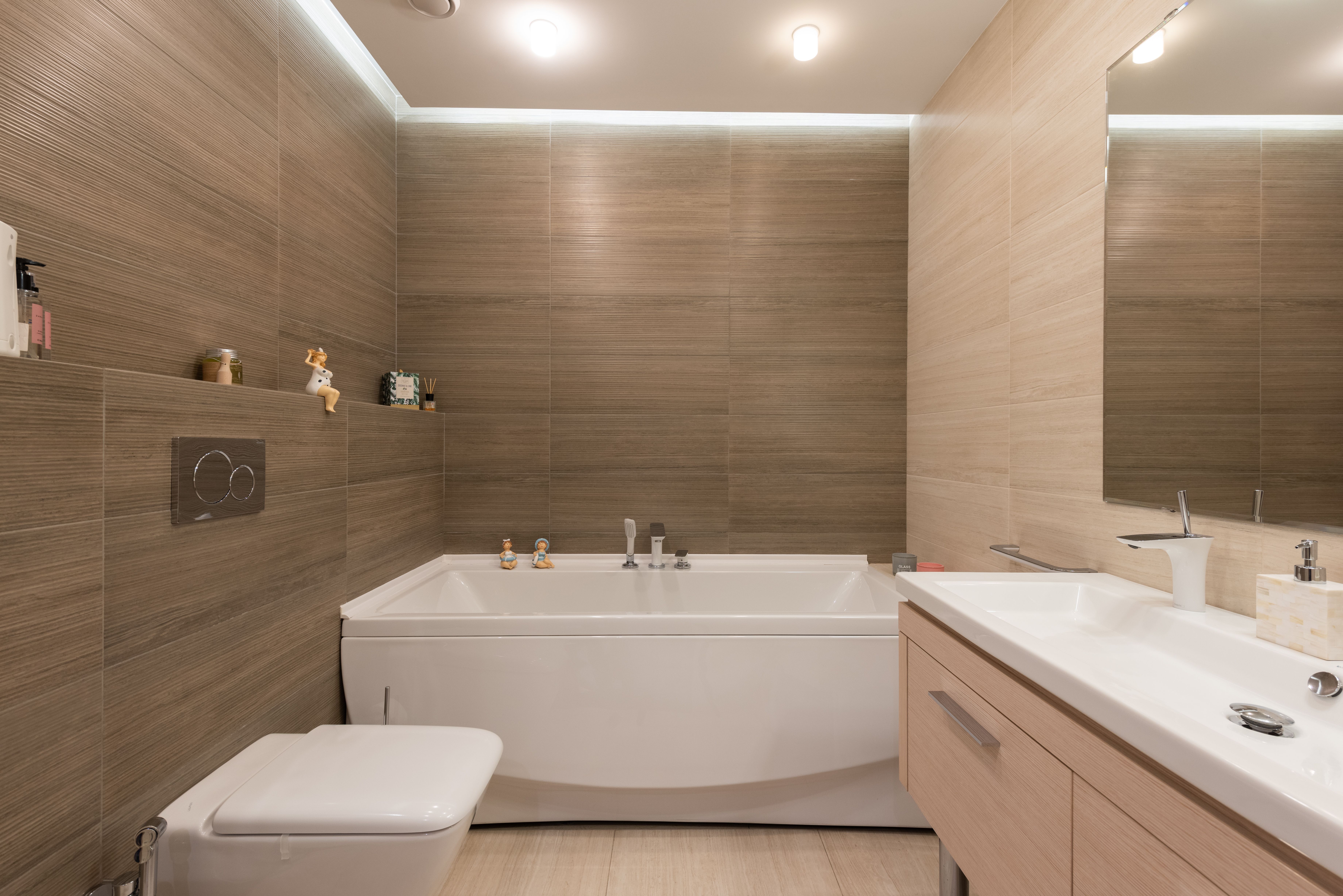 Modern bathroom with a white bathtub, toilet, wooden vanity, and beige tiled walls.