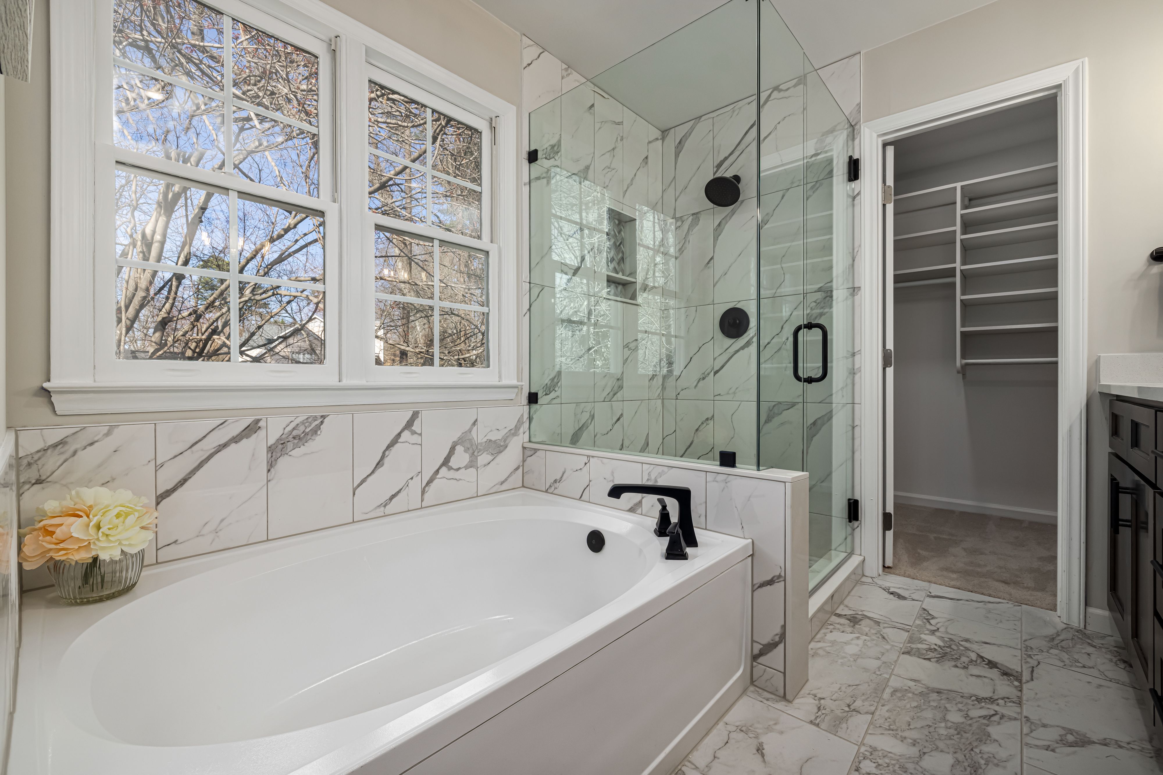 Modern bathroom with white tub, glass shower, marble tiles, window, and entry to walk-in closet.