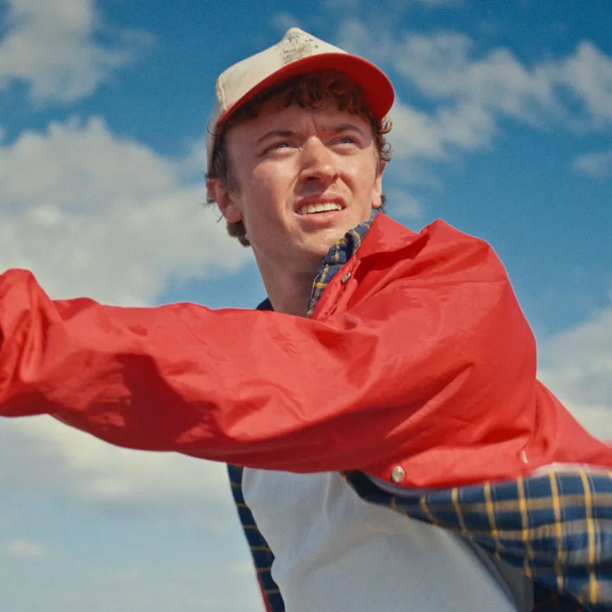 Young man in a red jacket and white cap looking into the distance against a blue sky with clouds.