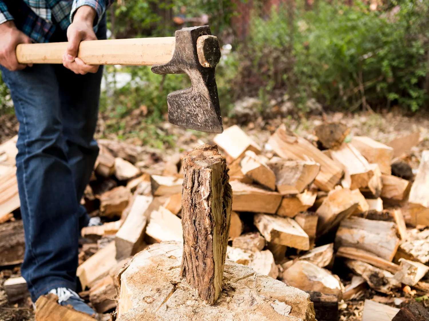 Person holding an axe poised above a log on a chopping block with a pile of split firewood in the background.