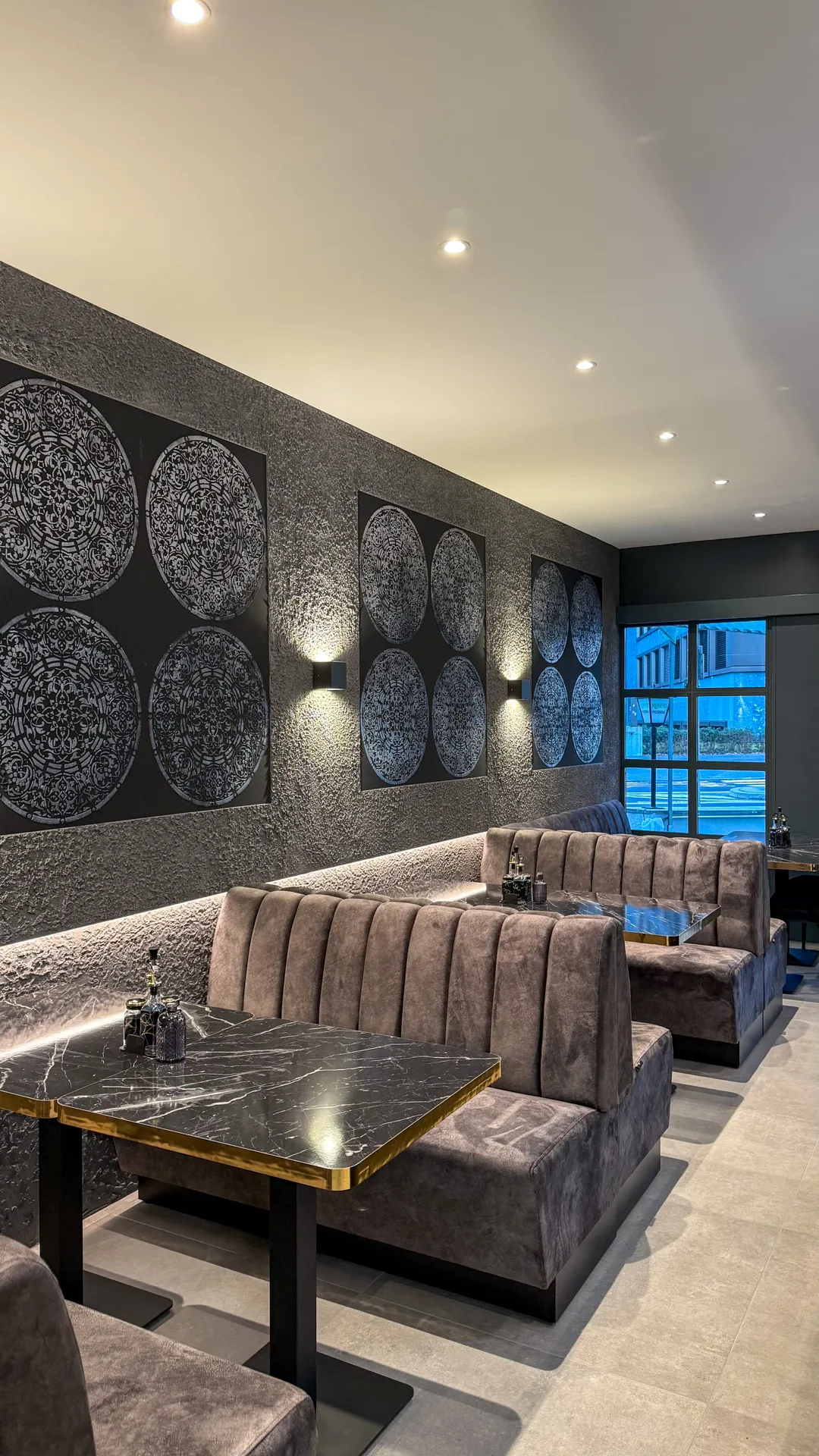 Modernes Restaurant mit grauen Polstersitzbänken, schwarzen Marmortischen und dekorativen runden Wandbildern an der strukturierten Wand.