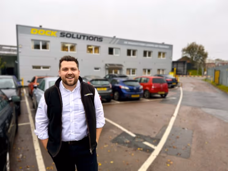 Man smiling with hands in pockets standing in a parking lot outside a building labeled 'Dock Solutions' with several parked cars.