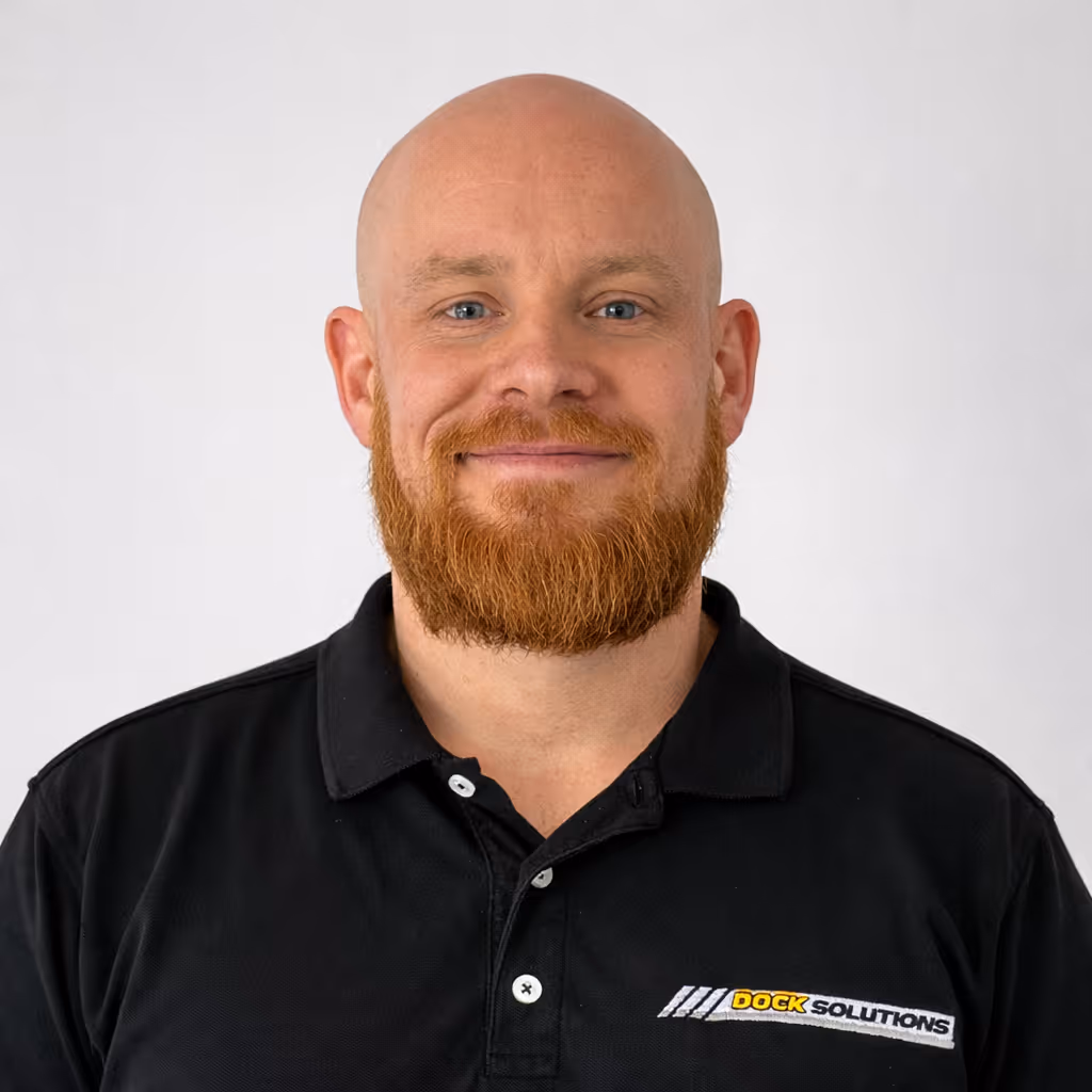 Smiling bald man with a red beard wearing a black polo shirt with a Dock Solutions logo on a plain white background.