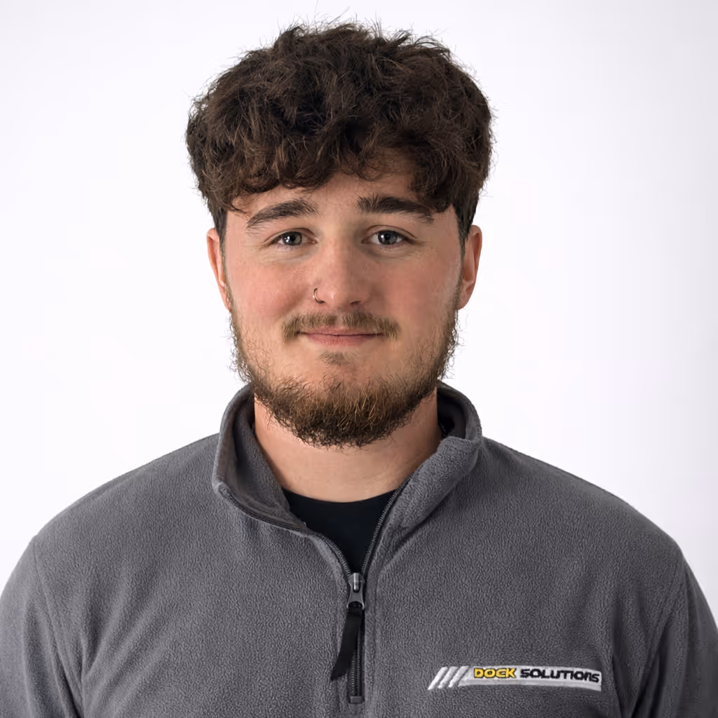 Young man with curly brown hair, beard, and nose ring wearing a gray fleece jacket with a Dock Solutions logo on the chest, smiling slightly against a plain white background.