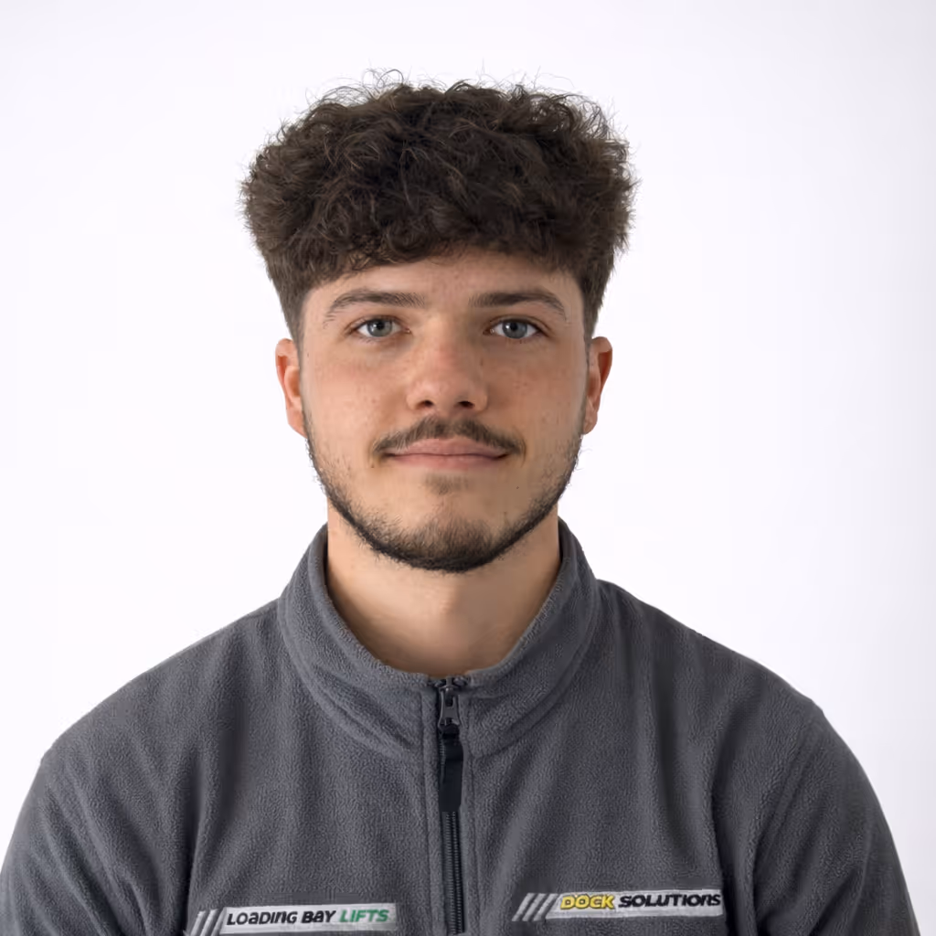 Portrait of a young man with curly hair and a beard, wearing a gray zip-up fleece jacket with a Loading Bay Lifts Dock Solutions logo, against a plain white background.