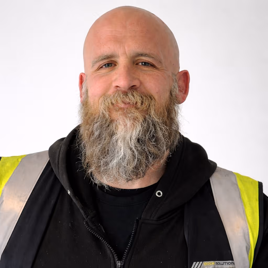Portrait of a bald man with a full graying beard wearing a black hoodie and a yellow safety vest against a plain white background.
