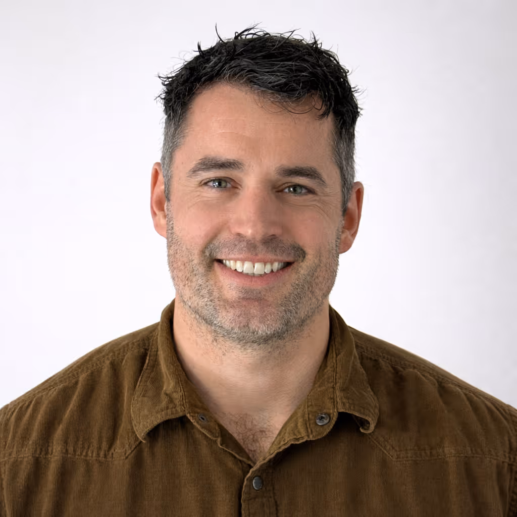 Smiling man with short, dark tousled hair and light stubble wearing a brown button-up shirt against a plain white background.