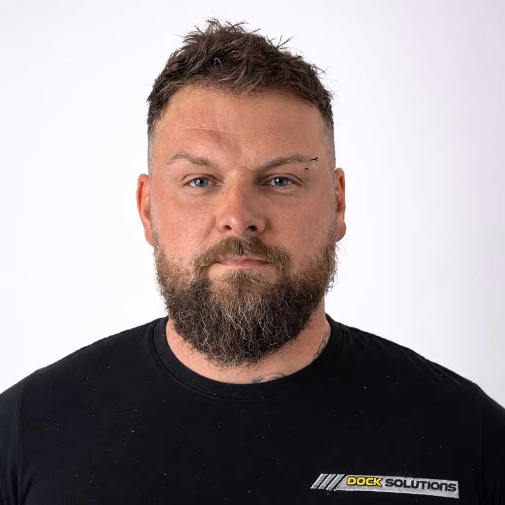 Portrait of a bearded man with short, spiked hair and an eyebrow piercing, wearing a black shirt with a Dock Solutions logo on the chest, against a plain white background.