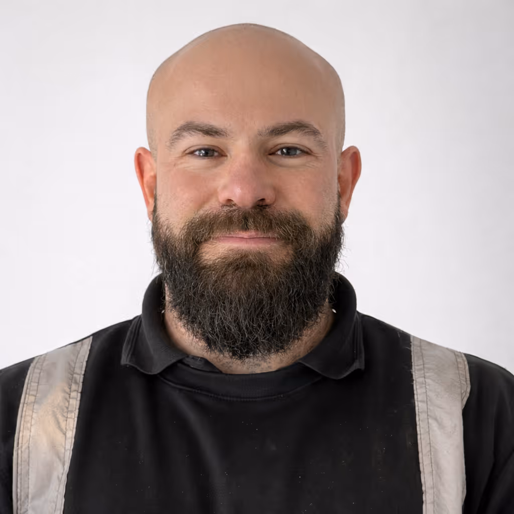 Portrait of a bald man with a full beard wearing a black shirt with beige suspenders, smiling slightly against a plain light background.