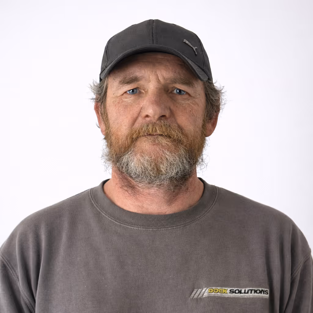 Middle-aged man with blue eyes and gray beard wearing a black Puma cap and gray sweatshirt with 'Dock Solutions' logo on the chest, facing forward against a plain white background.
