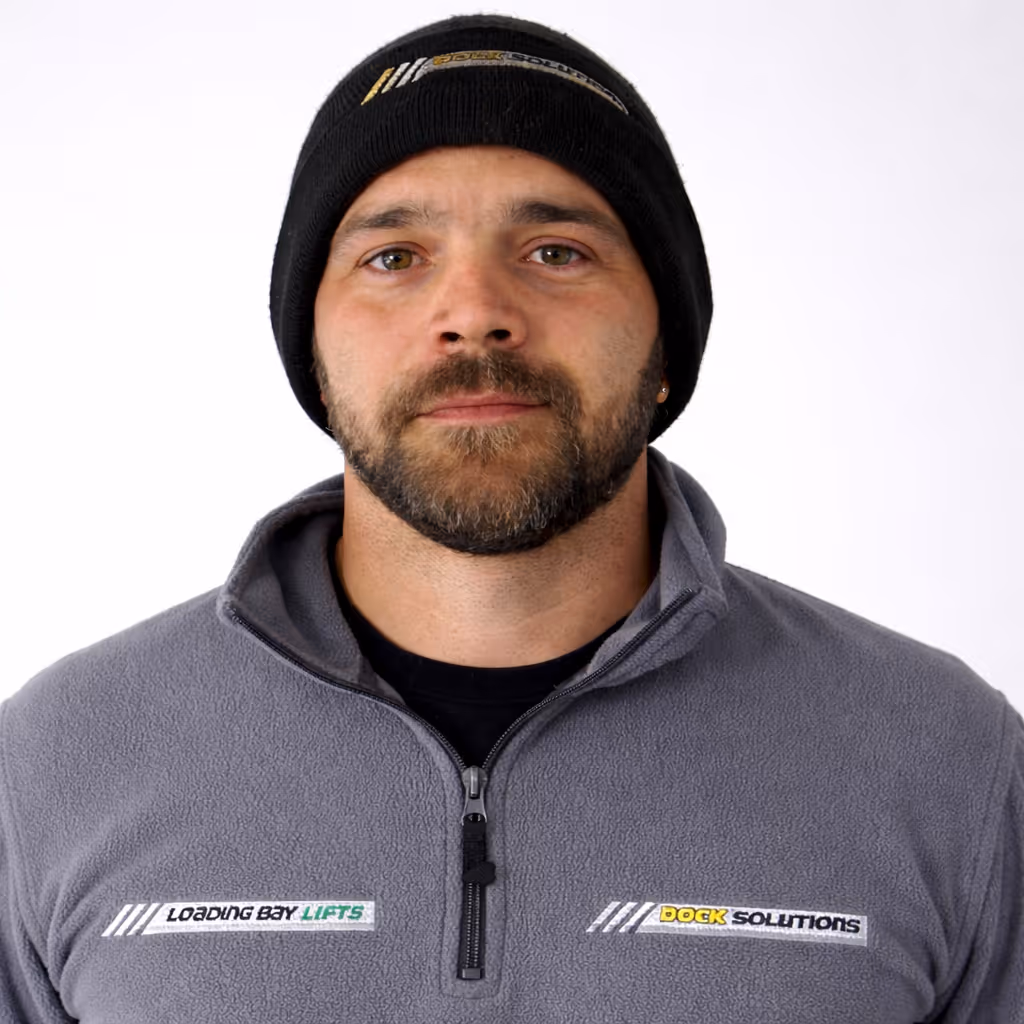 Man with a beard and mustache wearing a black beanie and a gray zip-up fleece jacket with logos reading 'Loading Bay Lifts' and 'Dock Solutions' on the front, standing against a white background.